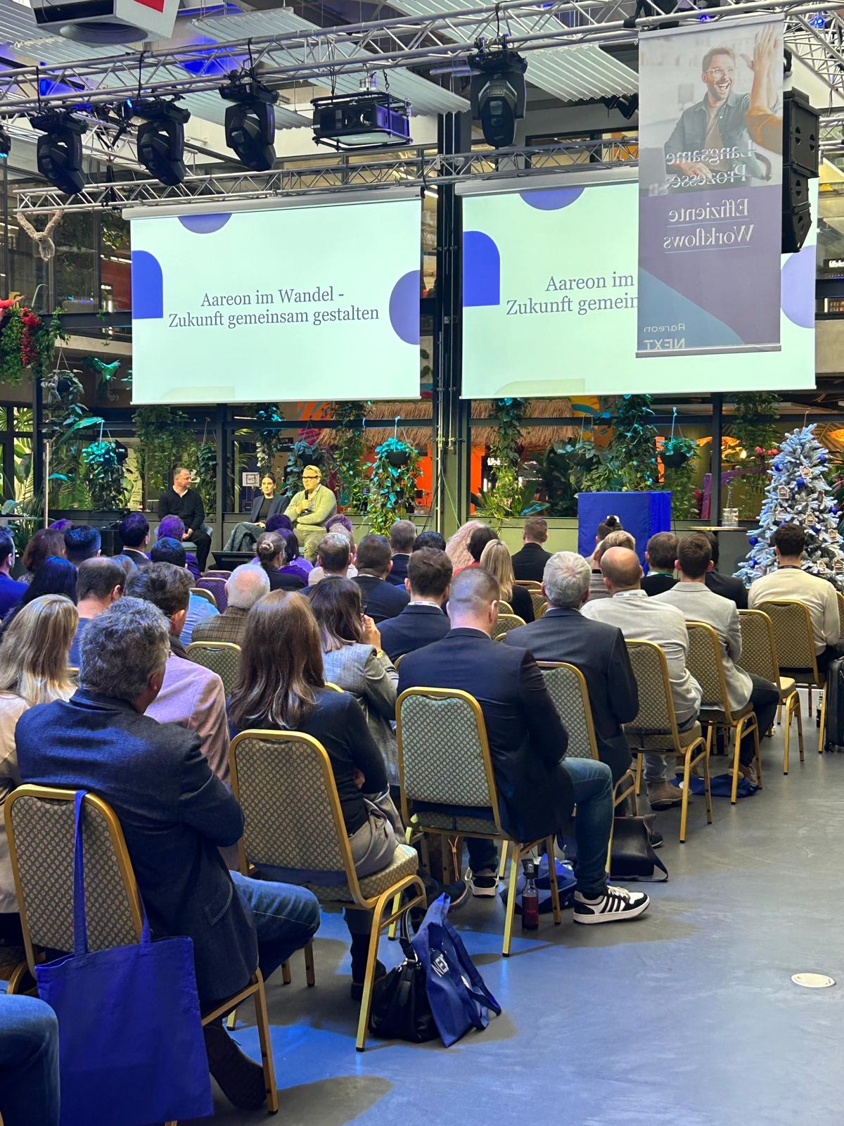 Bei der Eröffnungsmoderation der Veranstaltung sitzen 3 Personen auf der Bühne vor vielen Menschen im Plenum